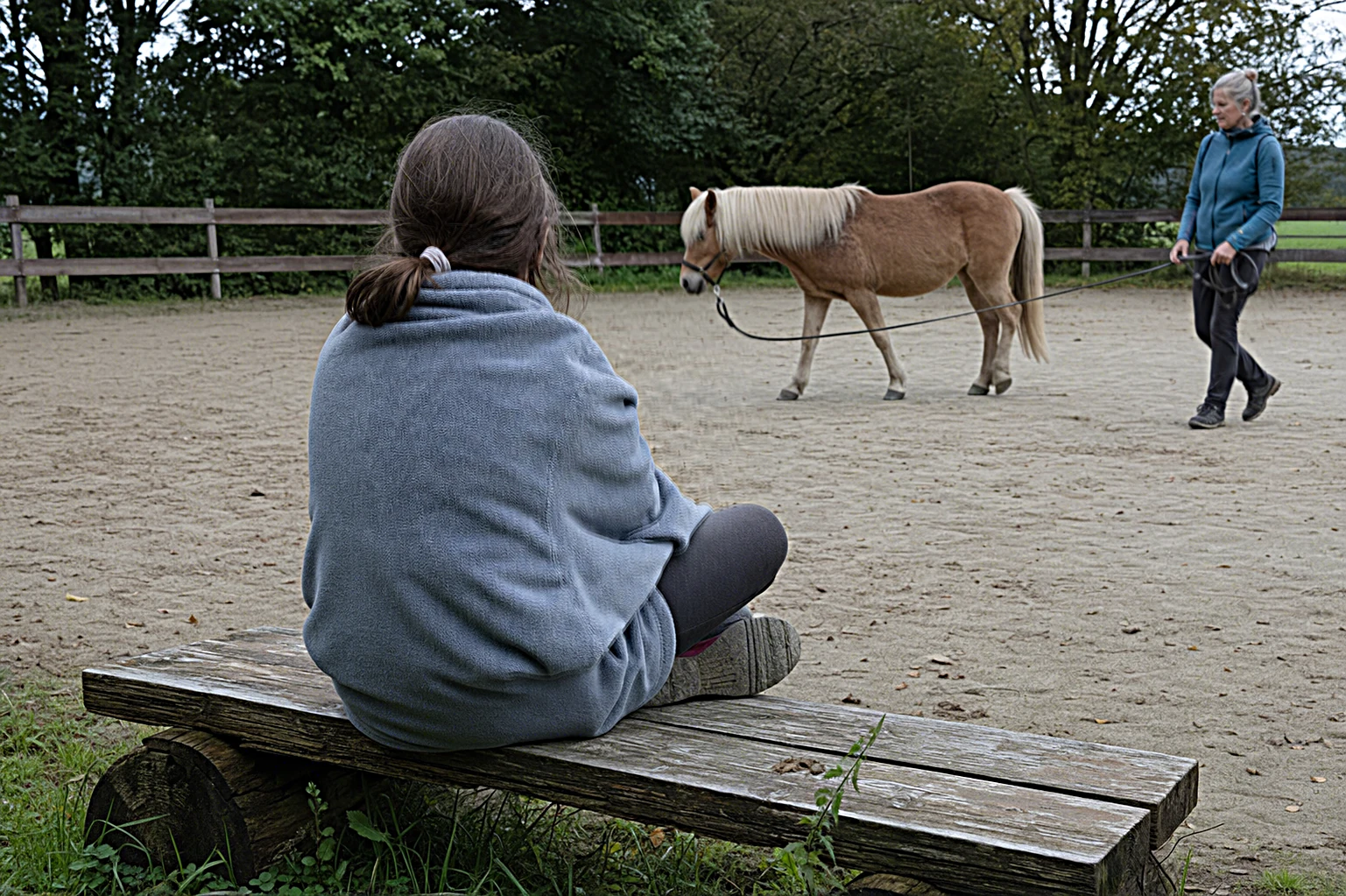 Kind im weichen Fleece-Plaid sitzt am Rand des Reitplatzes, im Hintergrund wird ein ruhiges Pony gefuehrt